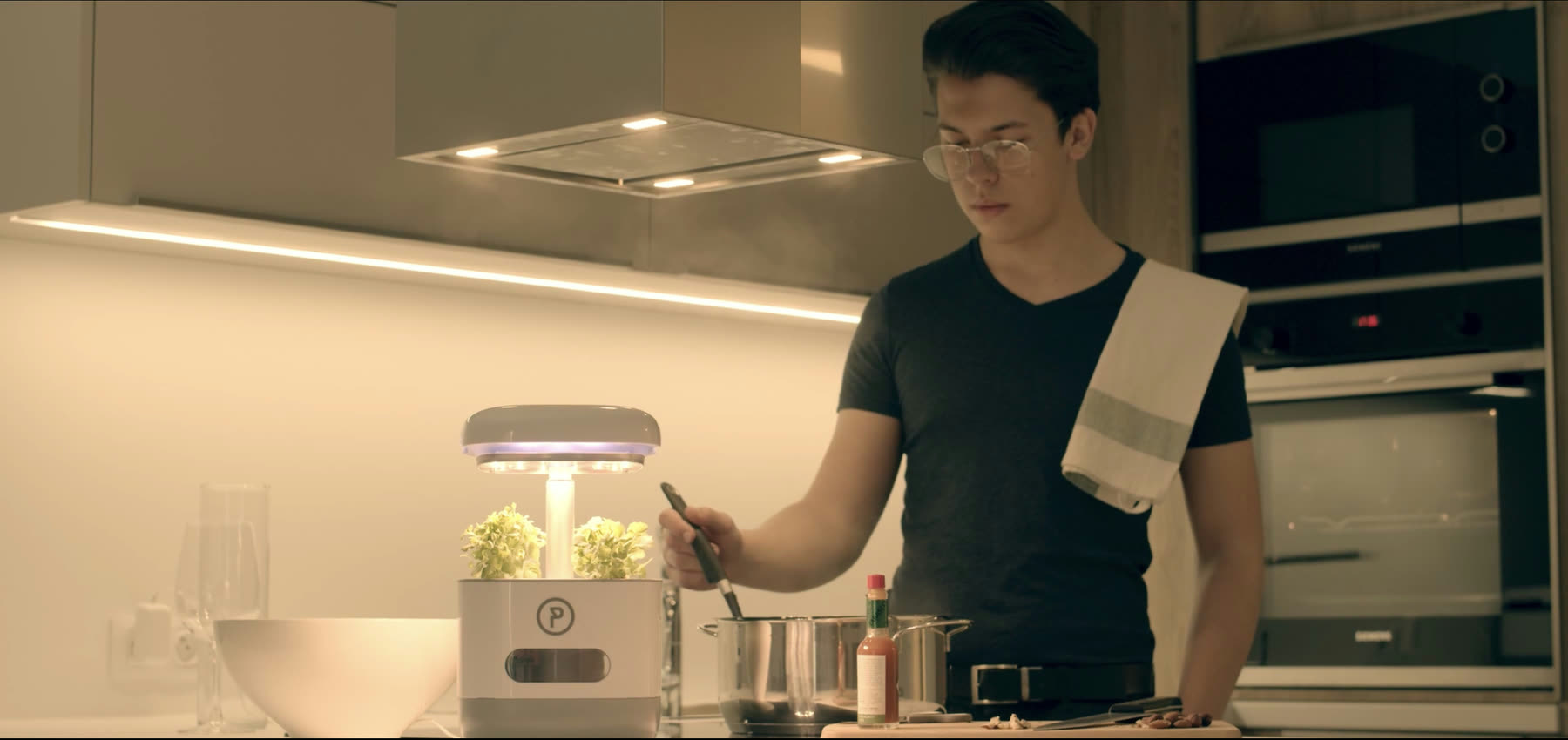 A young man cooking at a kitchen counter, the PonicTank beside him with its light ring glowing violet-white over young greens, a pan and chopping board in front of him.